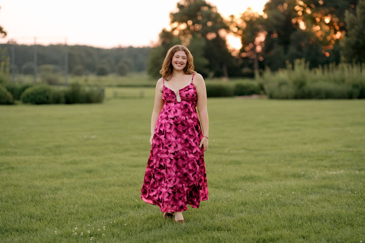 Young woman in pink floral maxi dress standing on lush green lawn at golden hour sunset.