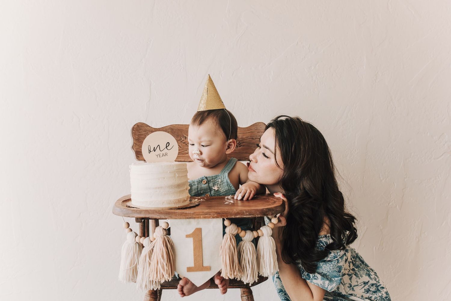 A mother blowing a kiss to her child who is sitting in a high chair with a birthday cake and party hat, celebrating his first birthday