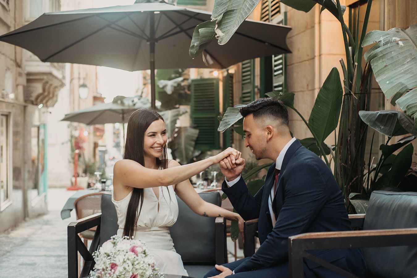 Bride and groom share a tender fist bump moment while seated at an outdoor cafe, surrounded by lush greenery.