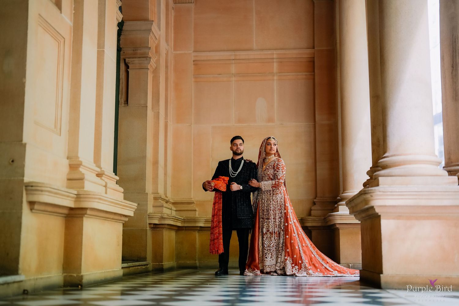 A couple poses elegantly in formal attire against classical stone columns in a grand architectural setting.