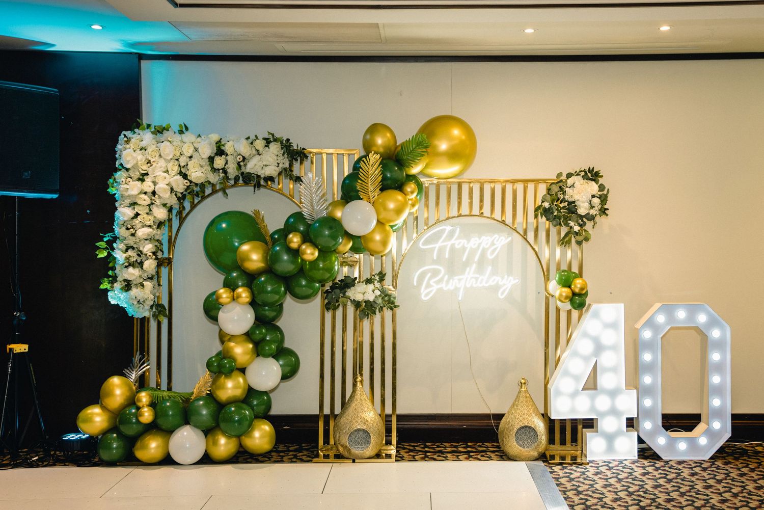 Elegant event backdrop with green and gold balloon garlands, floral arrangements, and illuminated numbers for decoration.