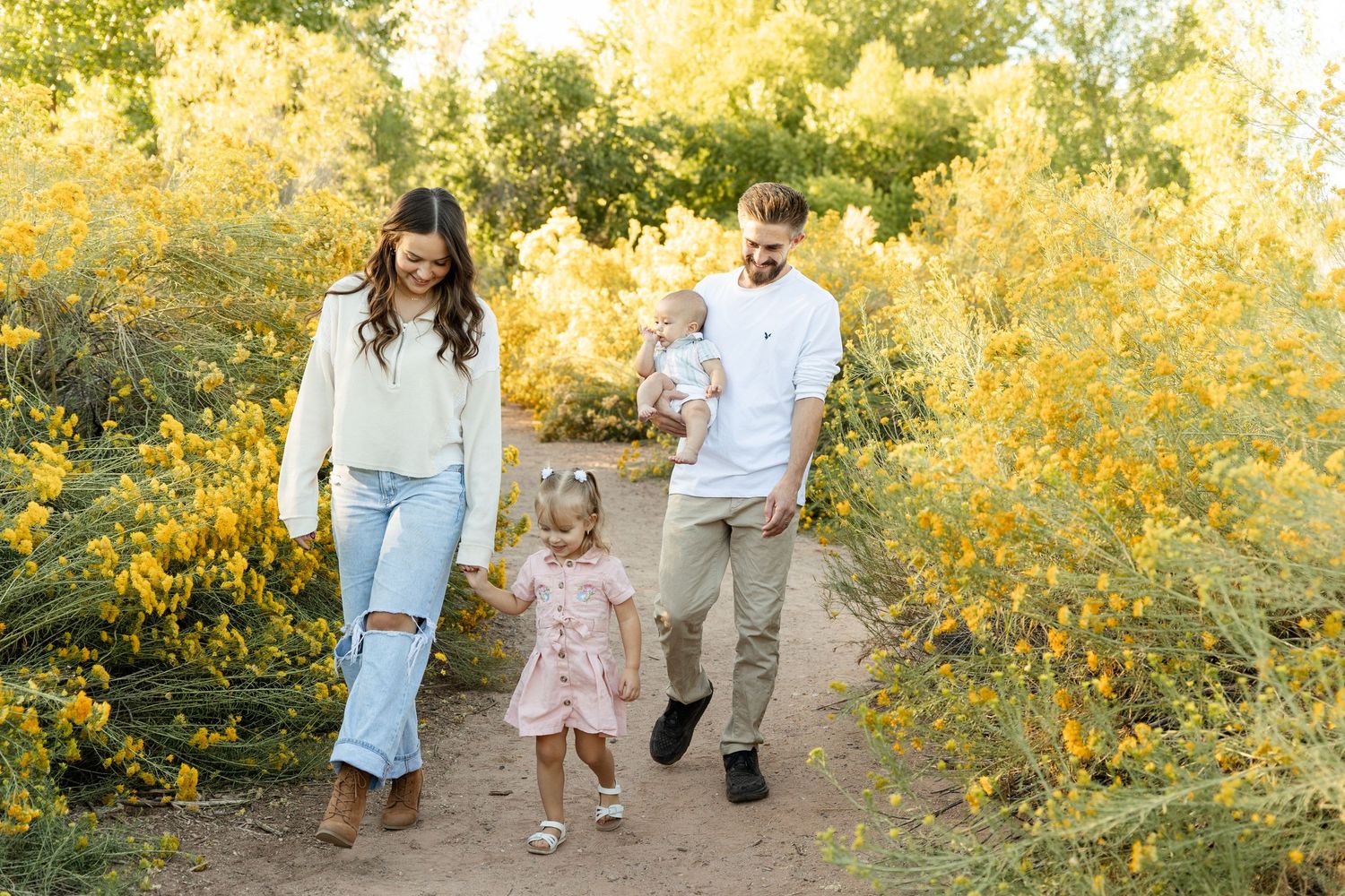Happy family with two young children walking on dirt path surrounded by vibrant yellow wildflowers.