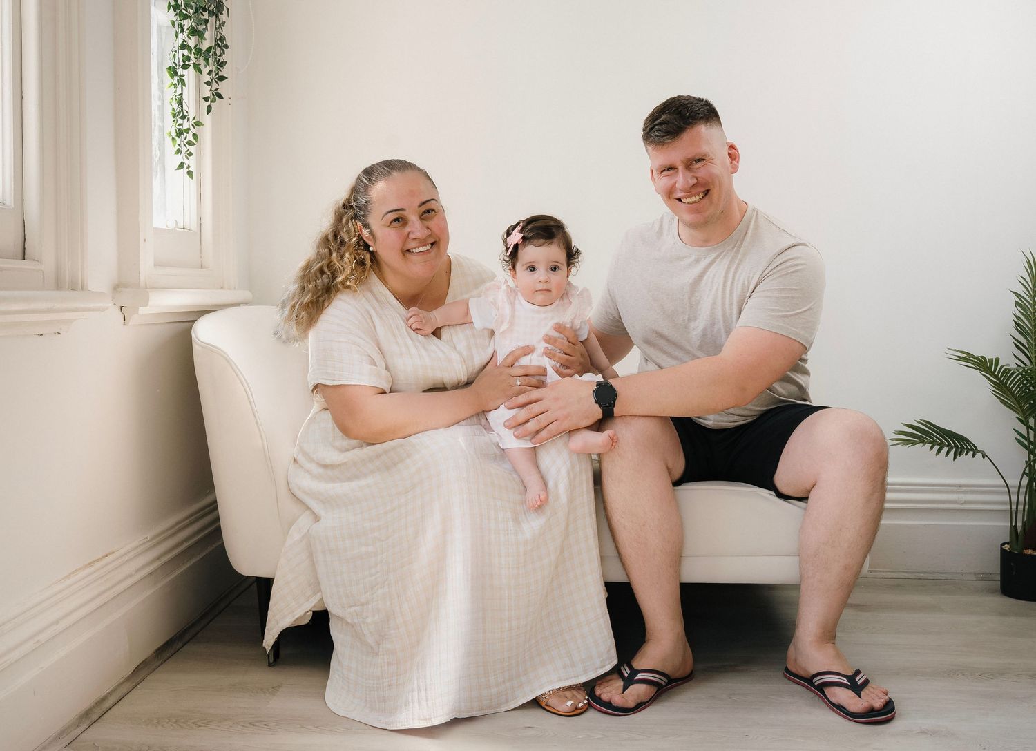 Happy family portrait with mother, father, and baby girl sitting on a white couch in a bright studio.