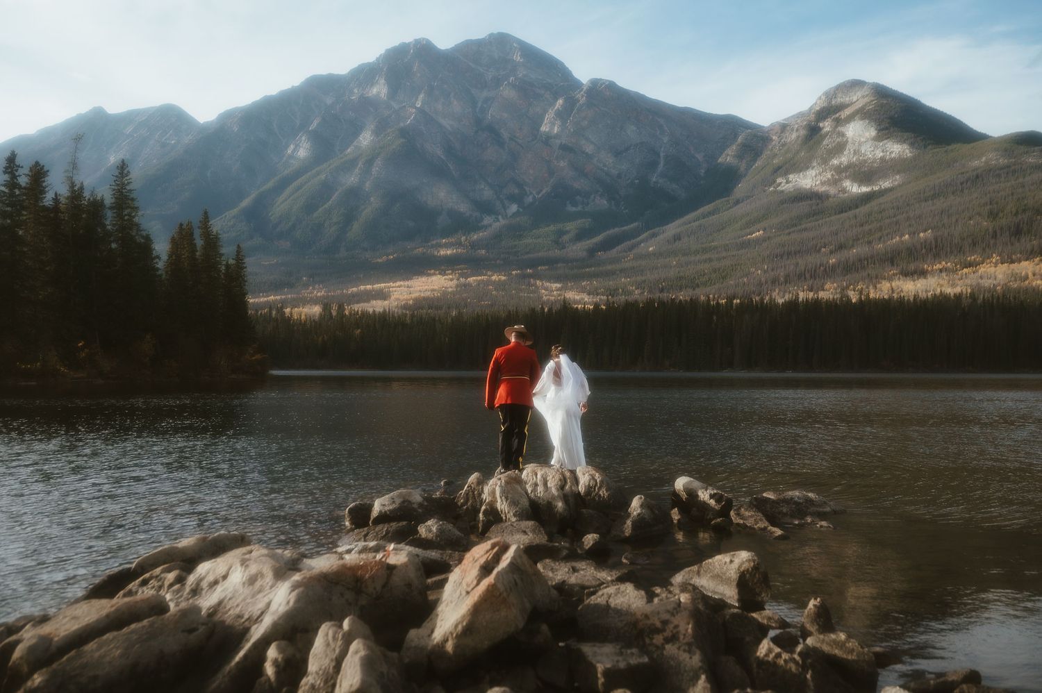A couple stands on rocky outcrop overlooking a serene mountain lake during their wedding day.