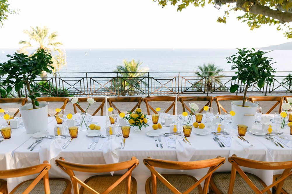 Rustic outdoor wedding reception setup with string lights and long wooden tables at sunset beneath olive trees.