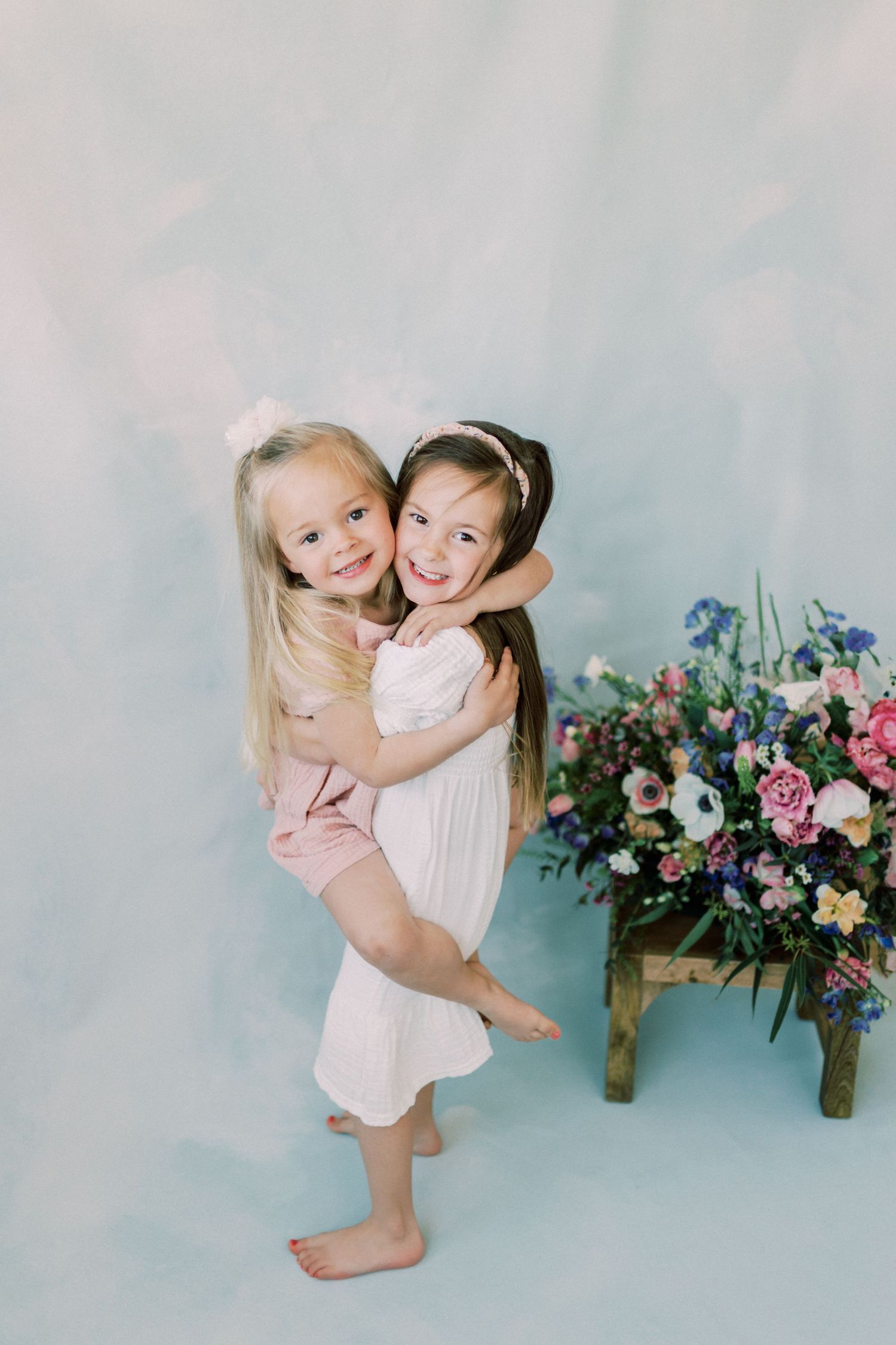 Two children in white and pink dresses sit together on a bench surrounded by floral arrangements in pink and green tones.