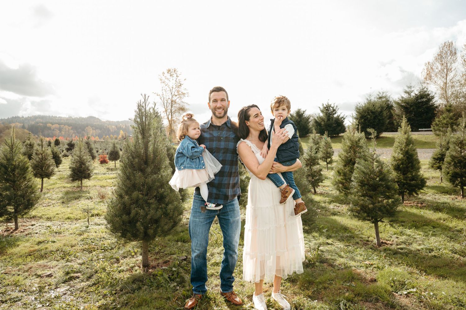 Couple walks through an evergreen tree farm on a sunny day wearing white and beige outfits.