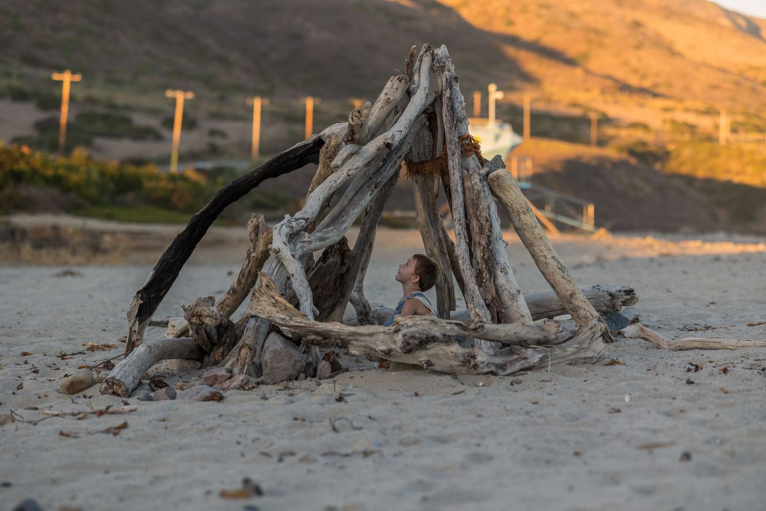 A beach campfire setup with driftwood logs arranged in a teepee formation at sunset against a mountainous backdrop.