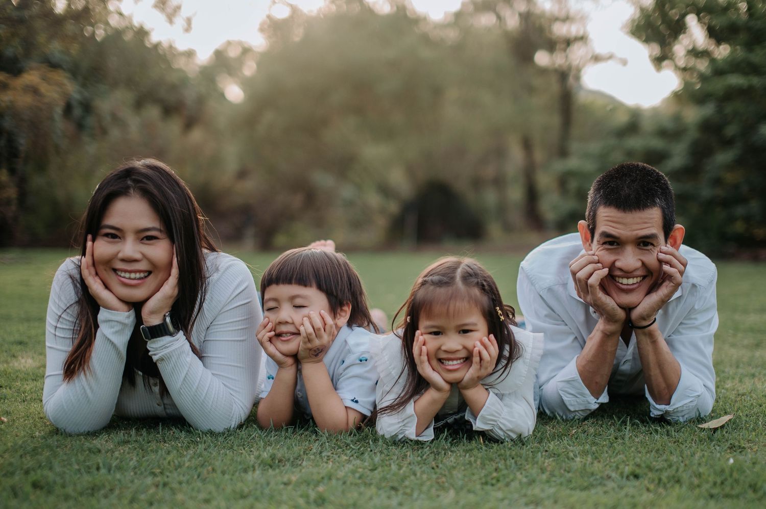 Family lays together on green grass in a park during sunset, all resting heads on hands while smiling at camera.