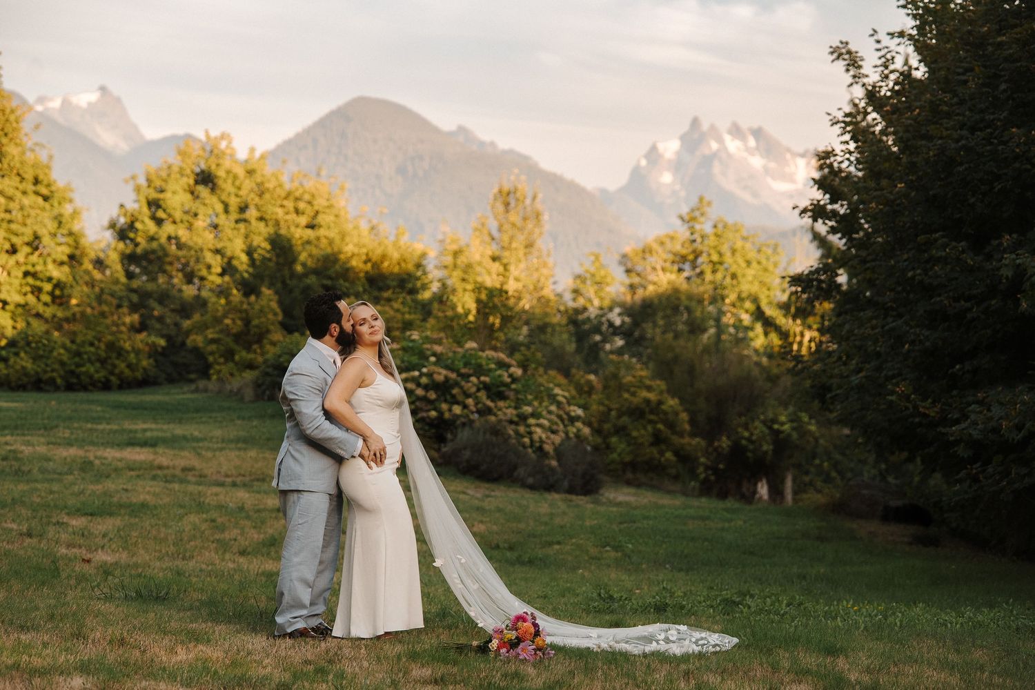 A couple embraces in a scenic mountain meadow during golden hour with mountains and trees in the background.