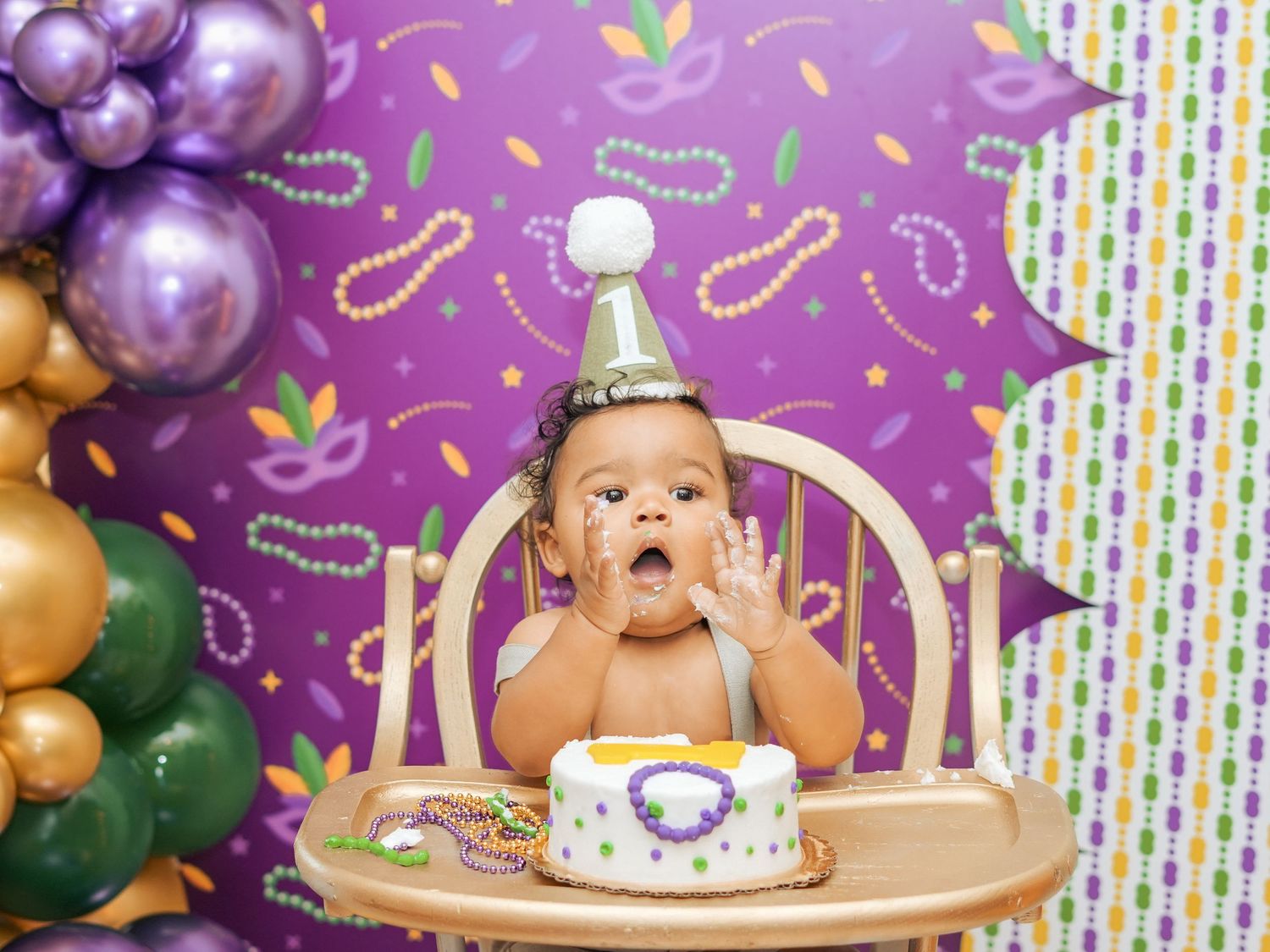 Baby in highchair enjoying first birthday cake with purple balloon decorations in background.