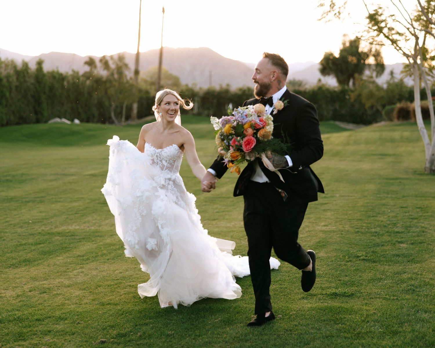A joyful bride and groom run hand in hand across a green lawn holding a colorful bridal bouquet.