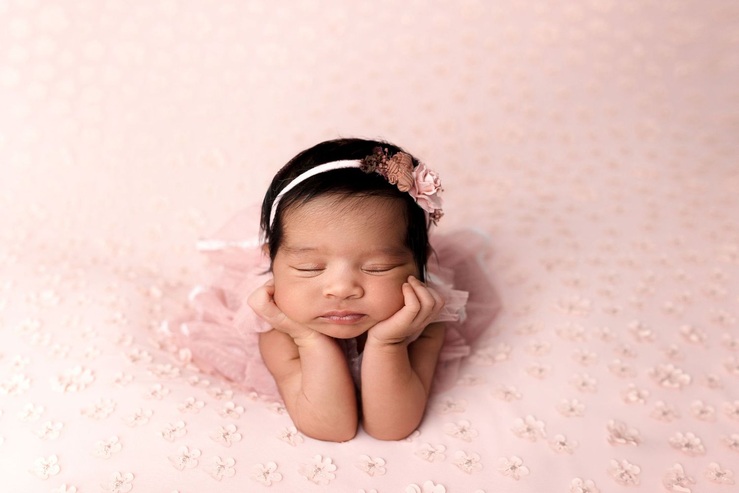 Newborn peacefully resting on pink blanket wearing floral headband in studio portrait setting.