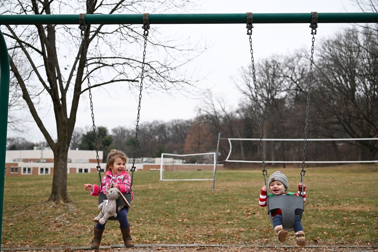 Two young children, a boy and a girl, are sitting in swings laughing while facing the camera. It is winter and they are bundled up in coats