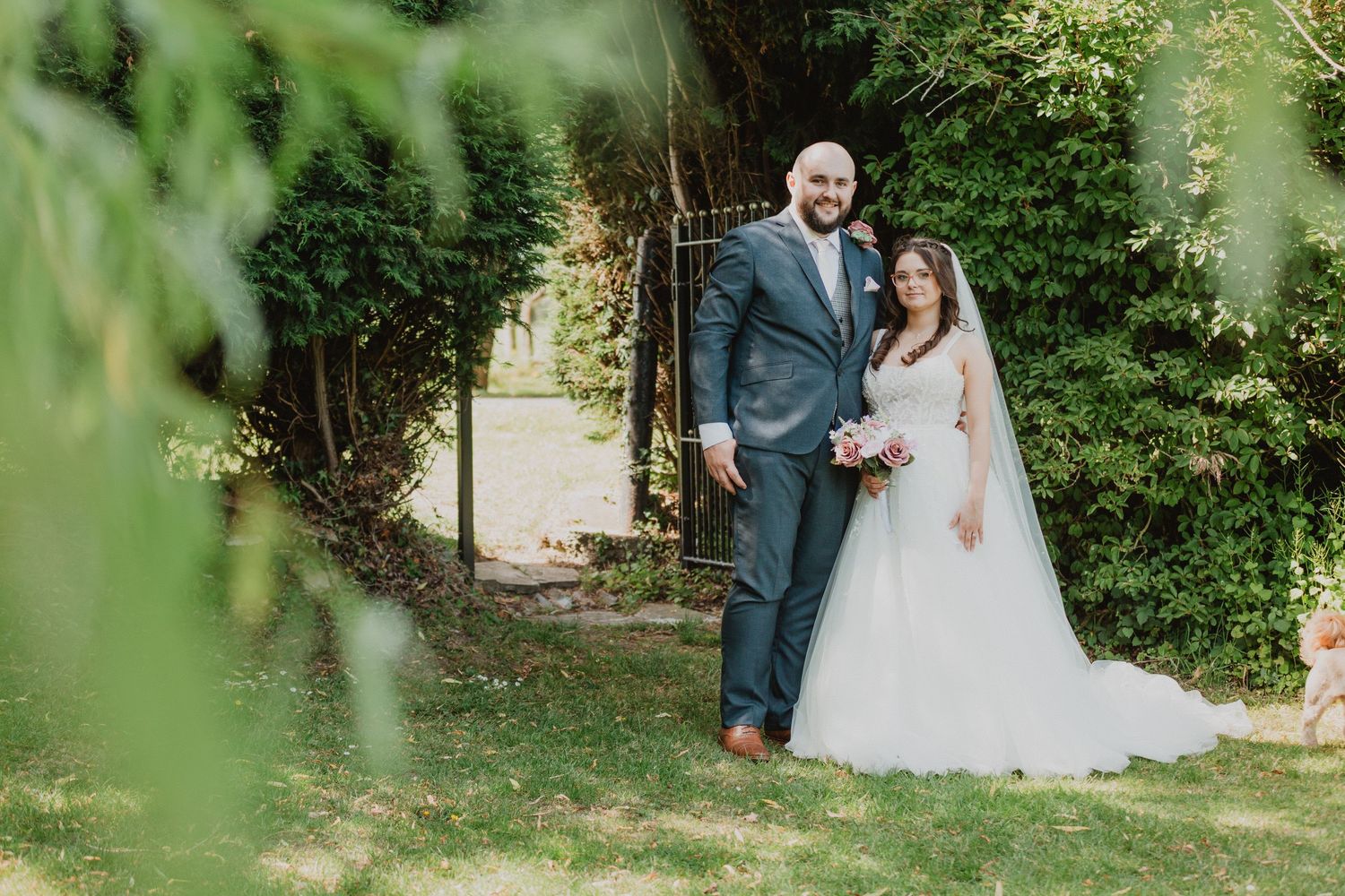 Bride and groom pose together in a lush garden setting, with the bride in a white gown holding a pink bouquet.