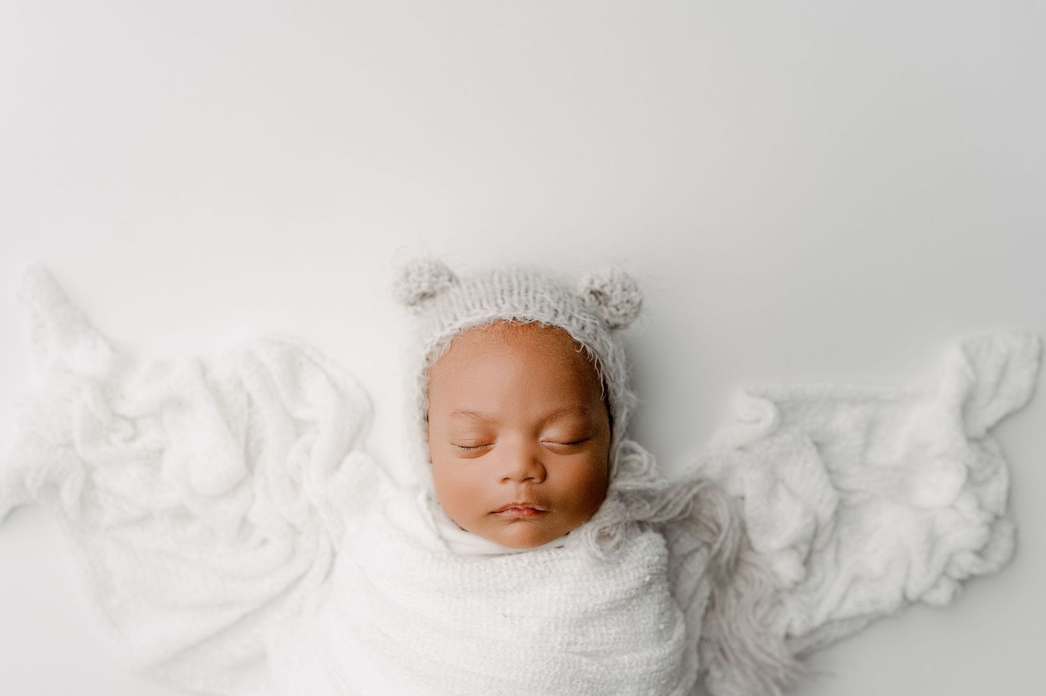 A peacefully sleeping newborn wearing a white bear hat lies on soft white fabric.