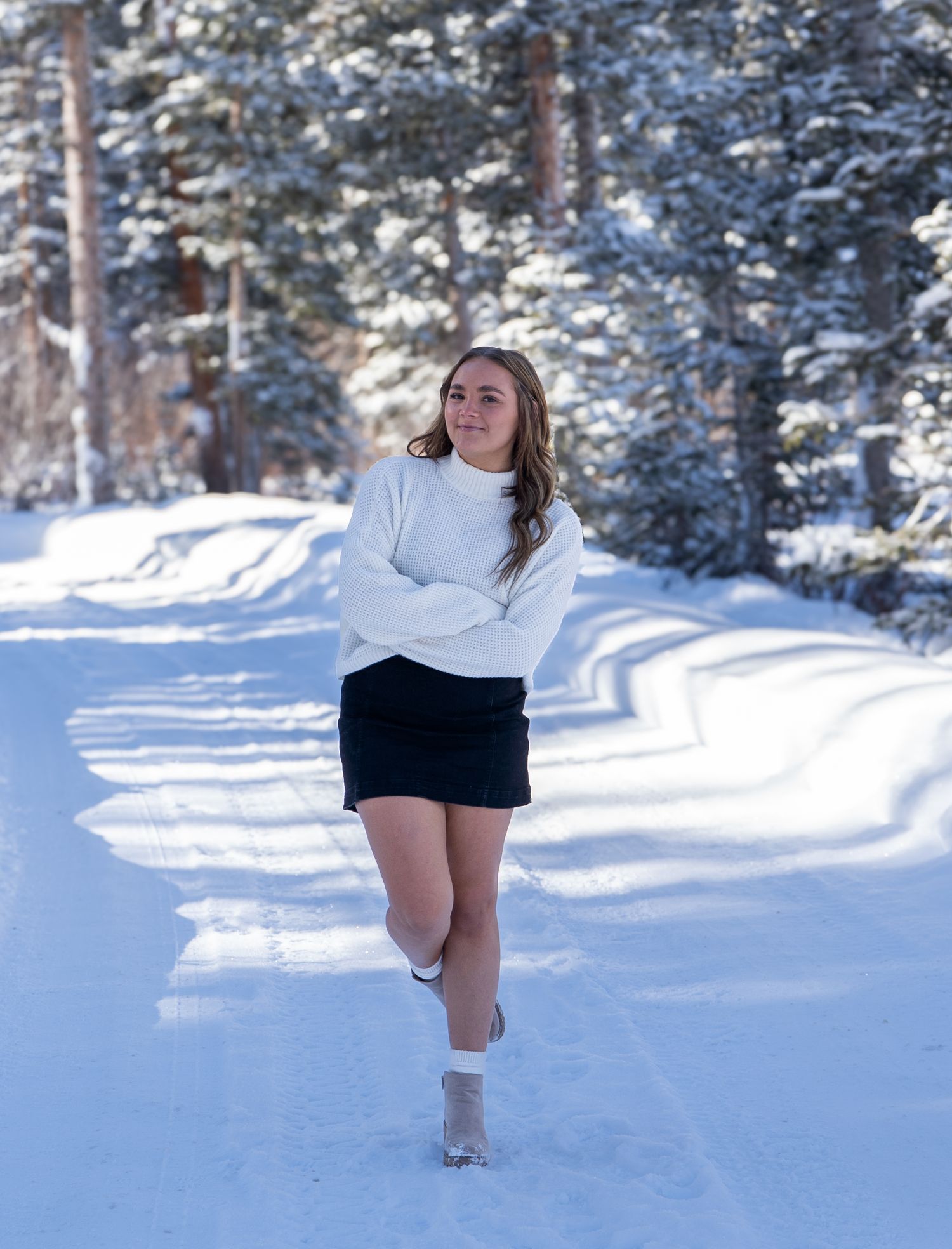 Person in white sweater and black skirt poses on snowy forest path during winter.