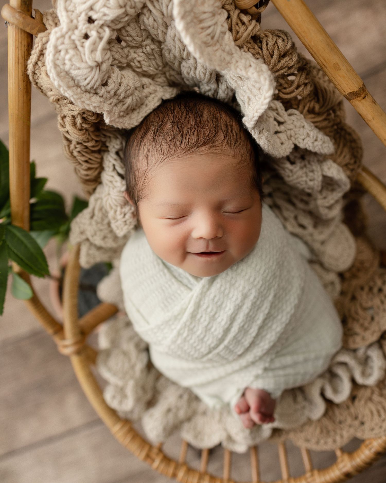 A sleeping newborn wrapped in a white knit blanket rests in a bamboo basket with plush textured bedding.