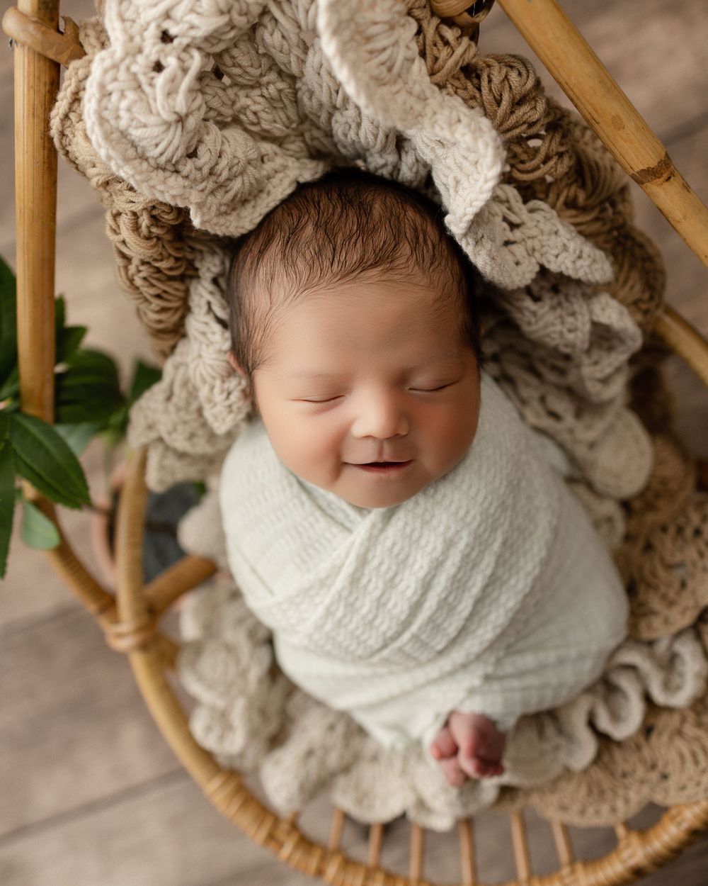A sleeping newborn wrapped in a white knit blanket rests in a bamboo basket with plush textured bedding.