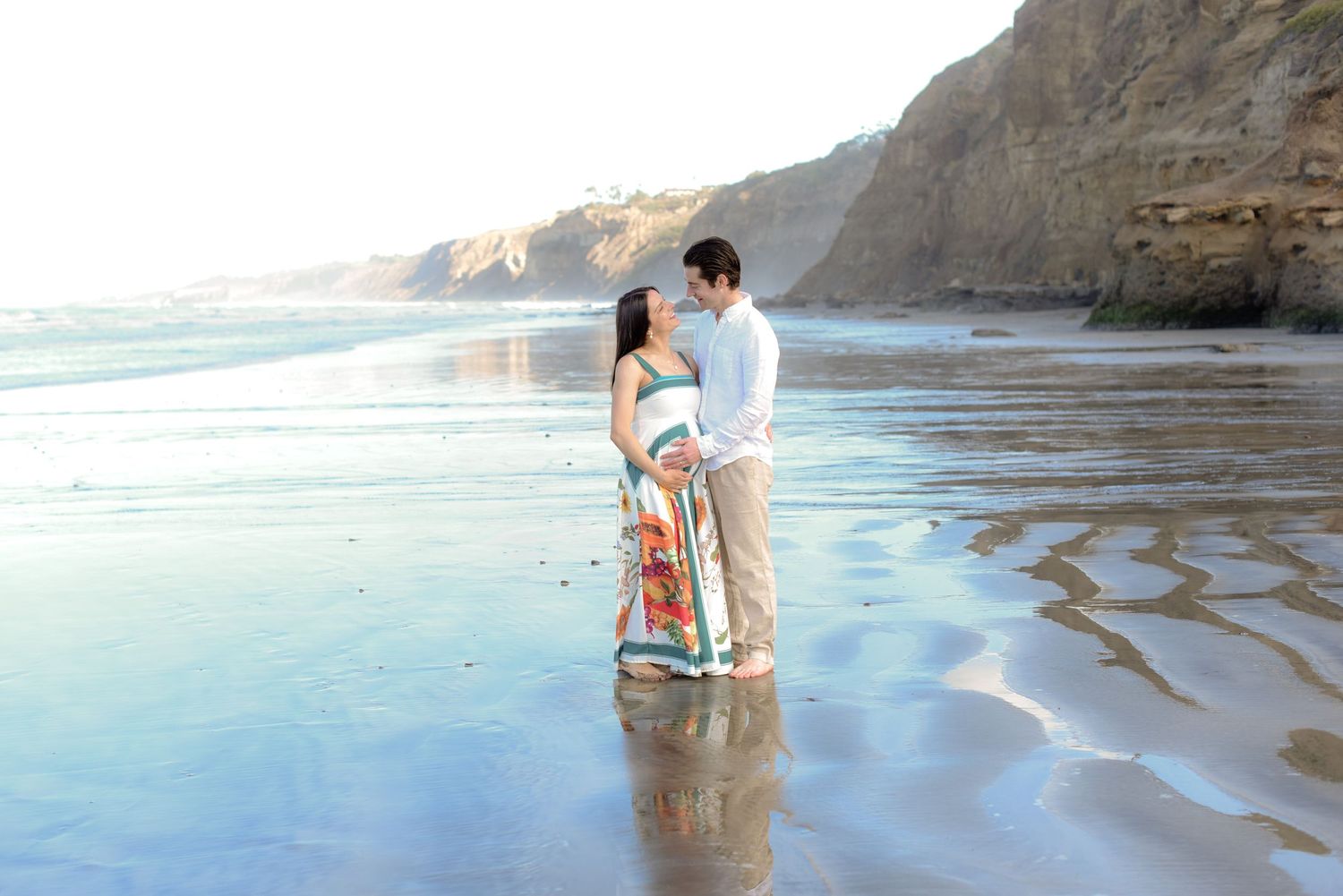 Couple standing together on reflective wet sand with coastal cliffs in background.