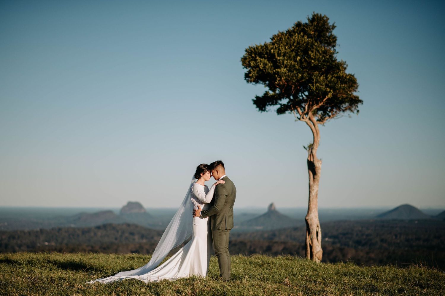 A couple embraces on a hilltop at sunset with a lone tree and mountain ranges in the background.