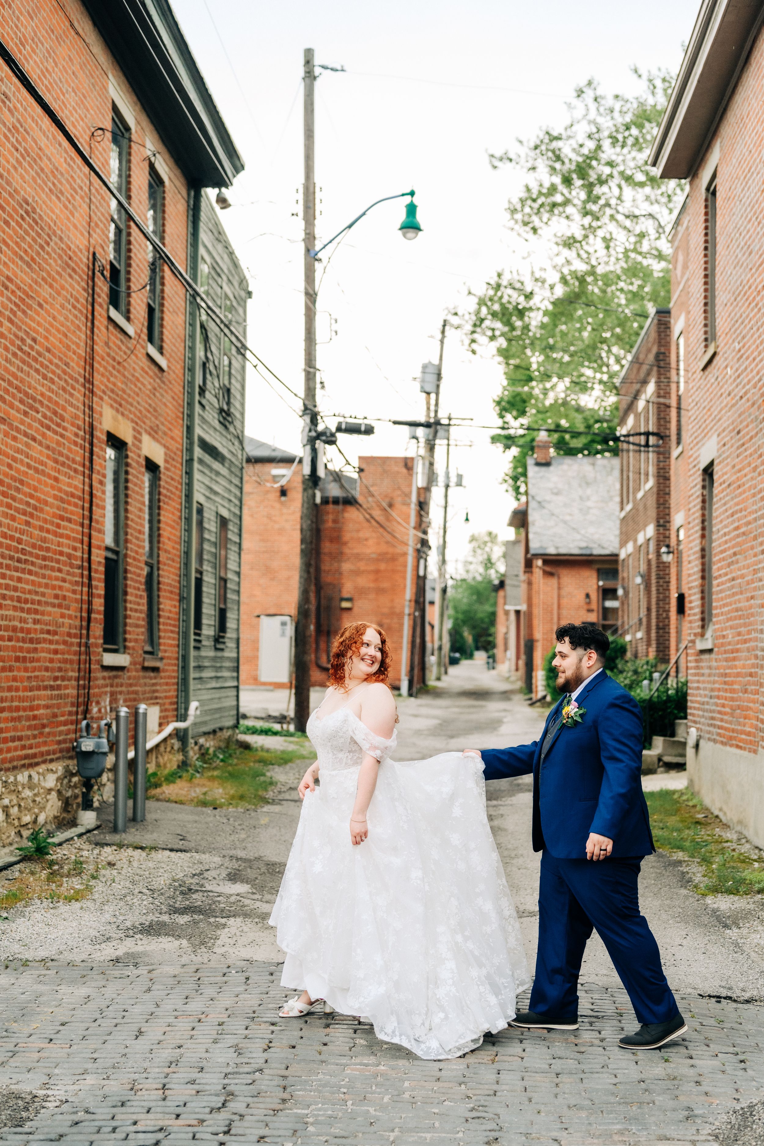 A couple in wedding attire share a joyful moment in an urban alley with brick buildings and cobblestone street.
