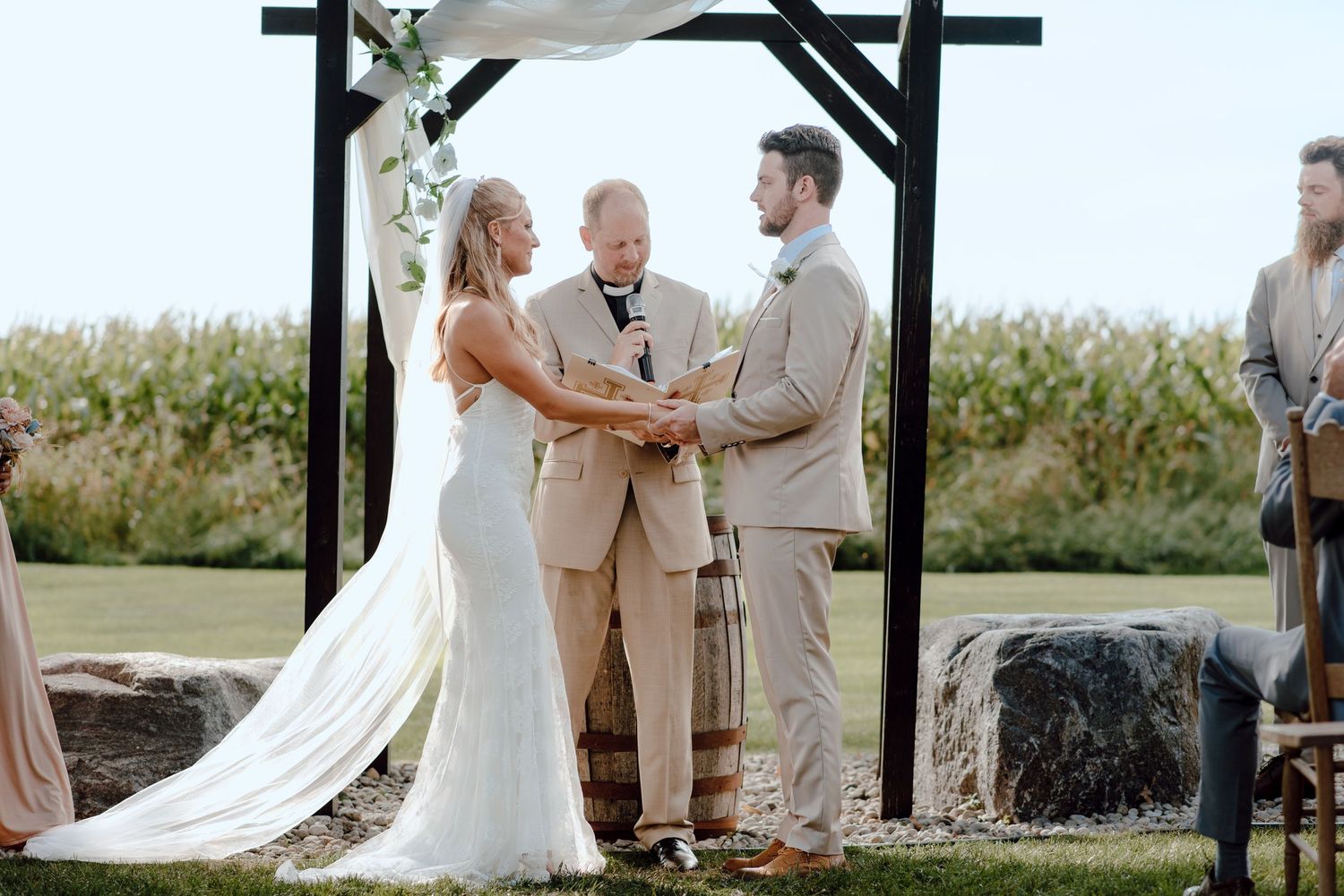 An outdoor wedding ceremony under a wooden arch with a cornfield backdrop.
