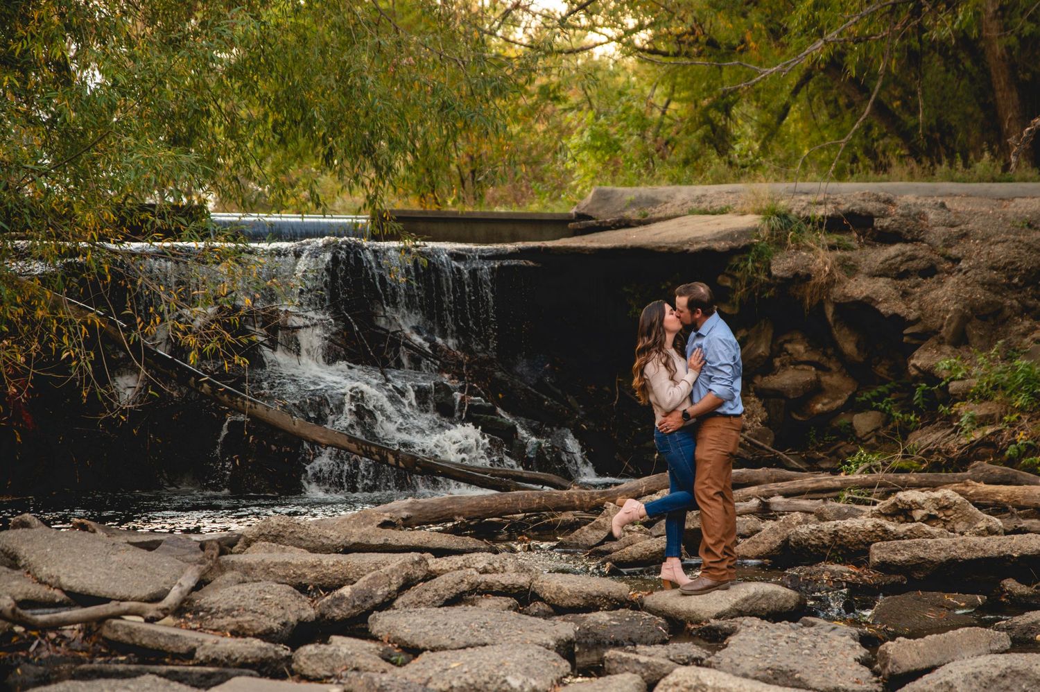 A couple embraces near a small waterfall on rocky terrain during golden hour.