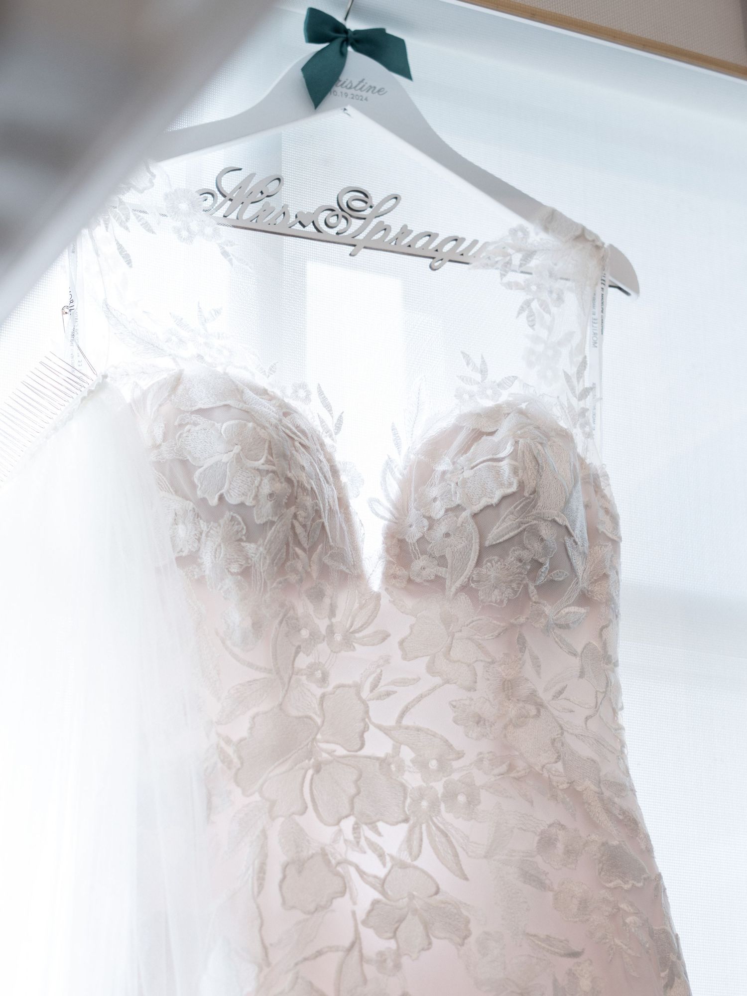 A delicate white lace wedding dress hangs on a hanger with a teal bow accent.