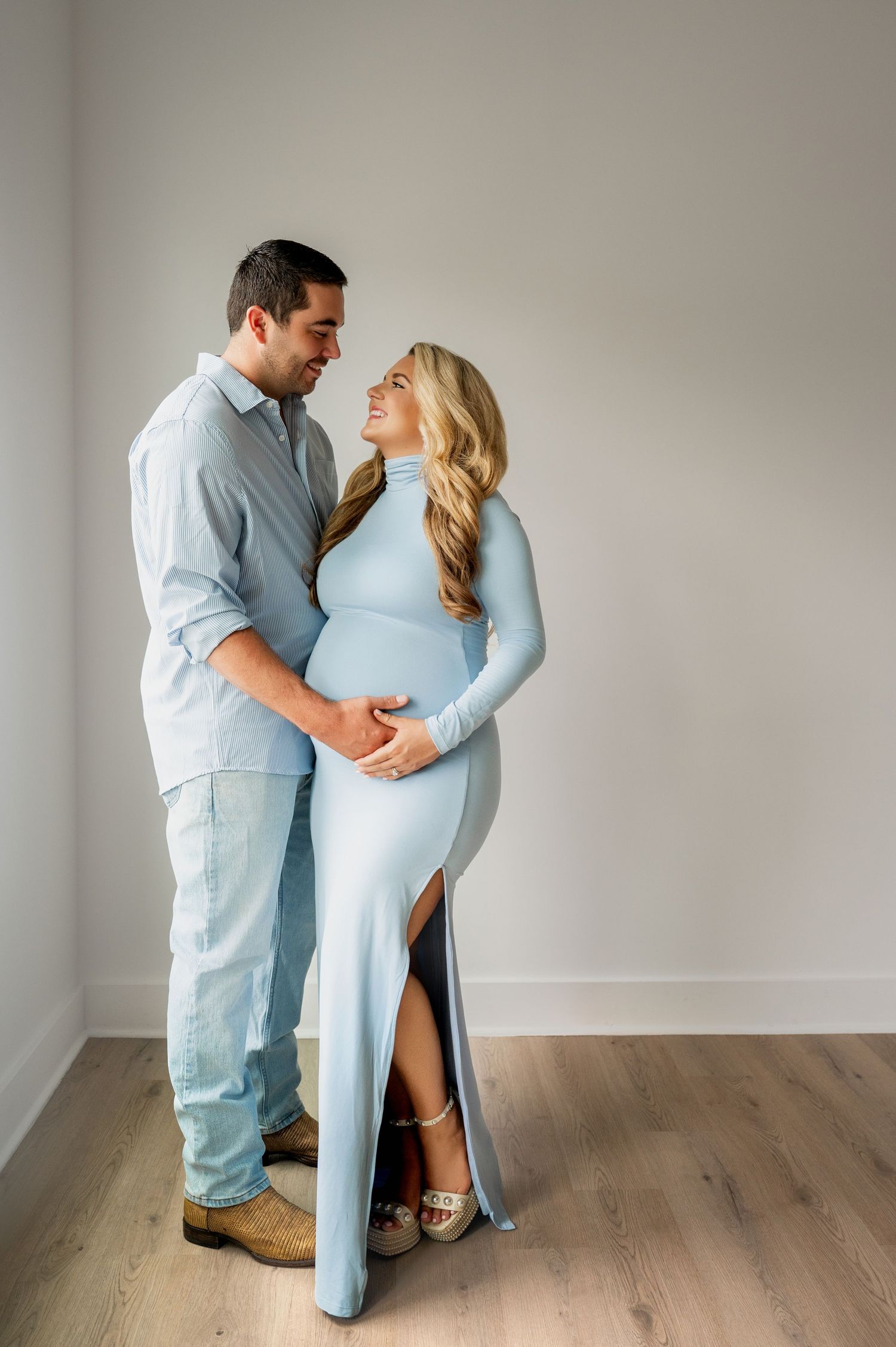 An expectant couple in matching blue outfits poses intimately against a neutral wall.