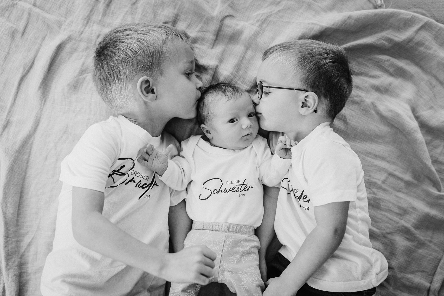 Die zwei "großen" Brüder küssen ihre kleine Babyschwester auf die Stirn links und rechts. Sie liegen zusammen auf dem Bett. Foto in S/W.