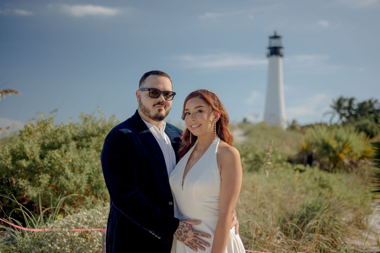 A couple in wedding attire embrace in a scenic meadow with a lighthouse in the background during golden hour.