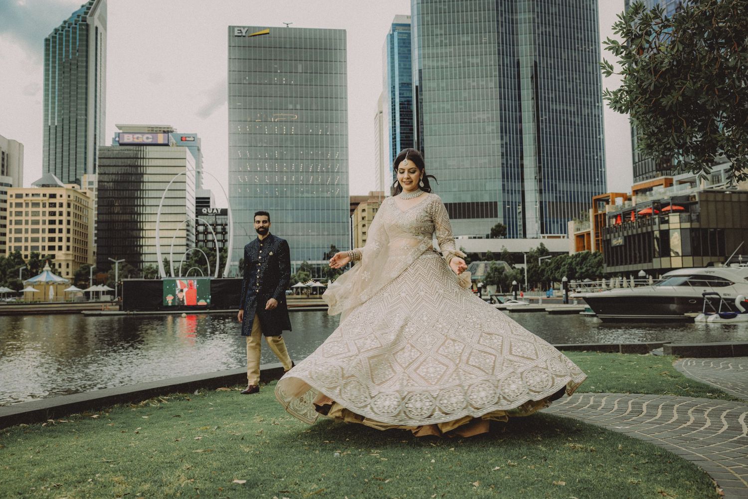 A bride in a white ballgown poses in front of modern glass skyscrapers and waterfront architecture in an urban setting.