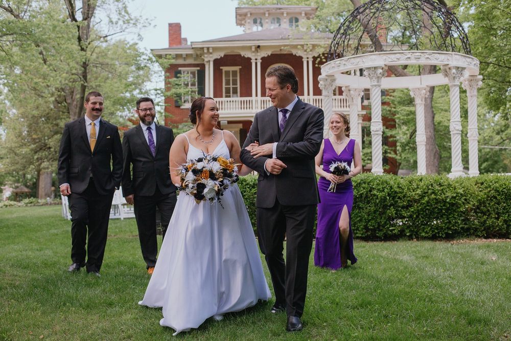 A smiling bride and groom walk in front of their wedding party in front of the Larimore venue.