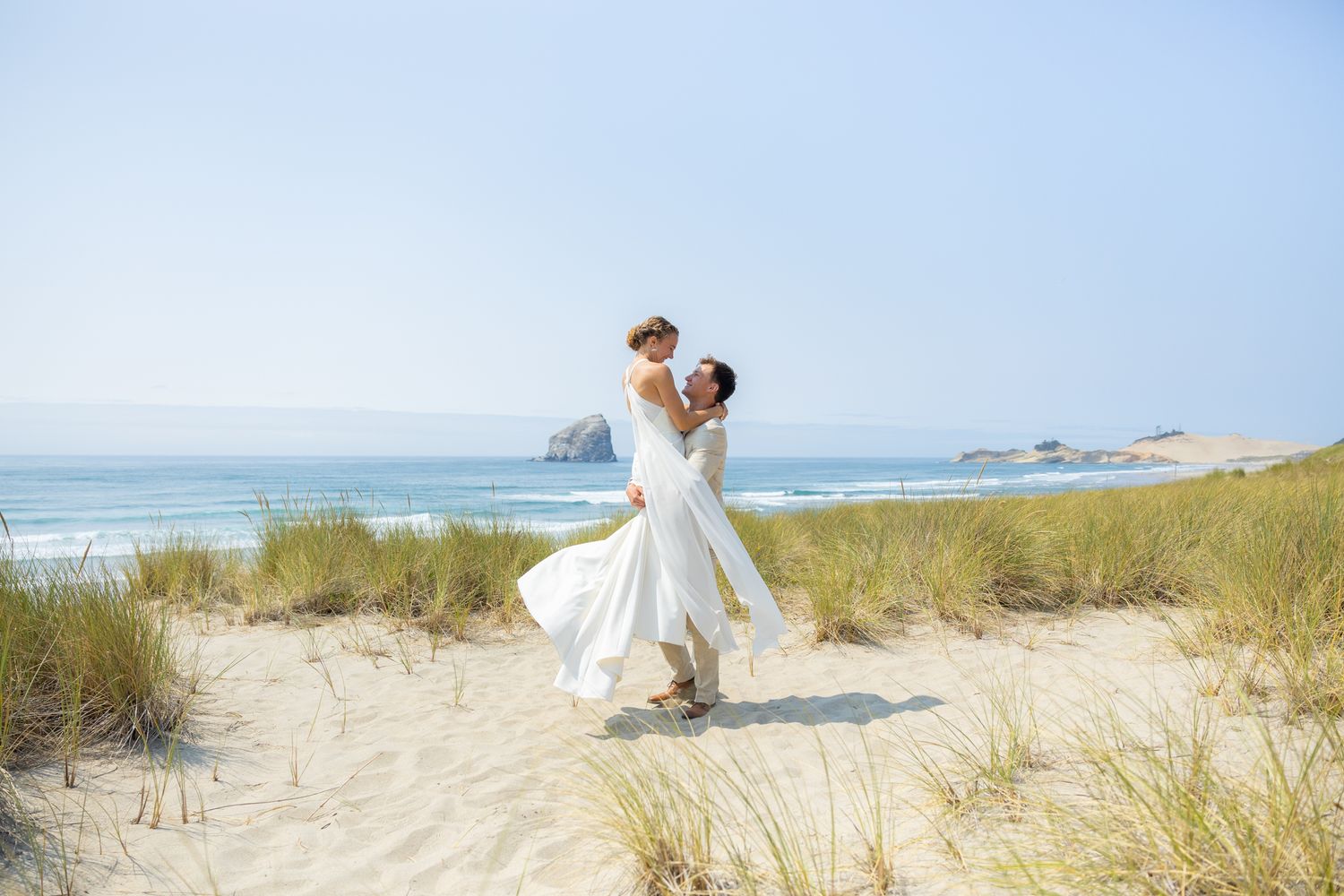 Couple in formal wedding attire embracing on a scenic beach with sand dunes and ocean views.