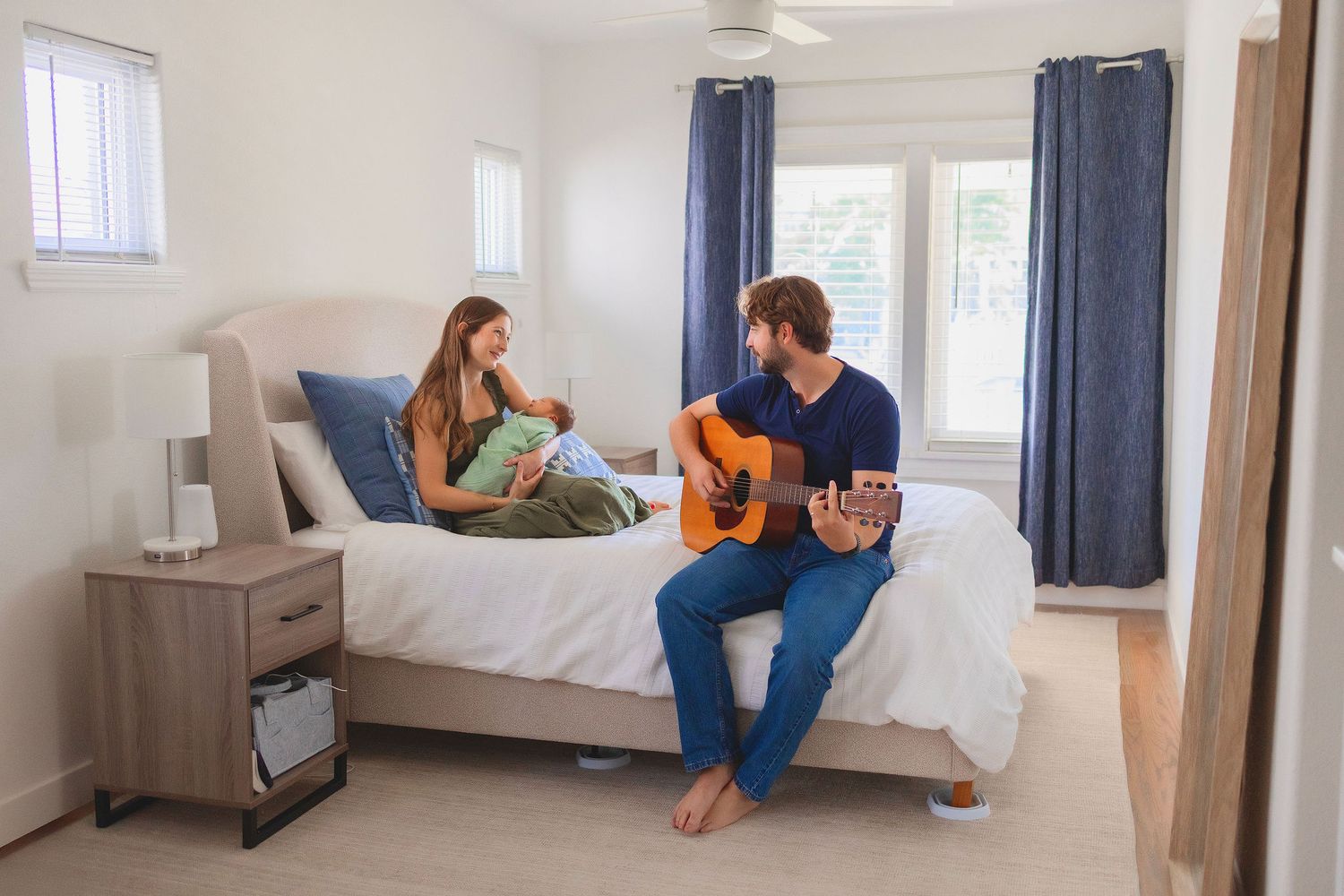 A couple, Mom and Dad, relaxes on a bed in a bright bedroom while Dad plays acoustic guitar and Mom smiles while holding newborn baby