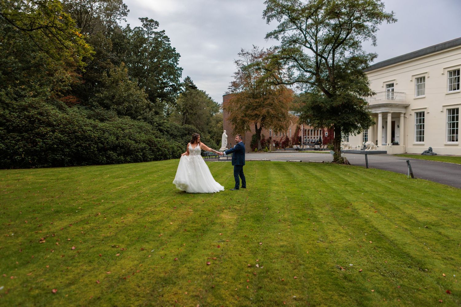 A couple in wedding attire stands on a lush green hillside next to a white building on an overcast day.