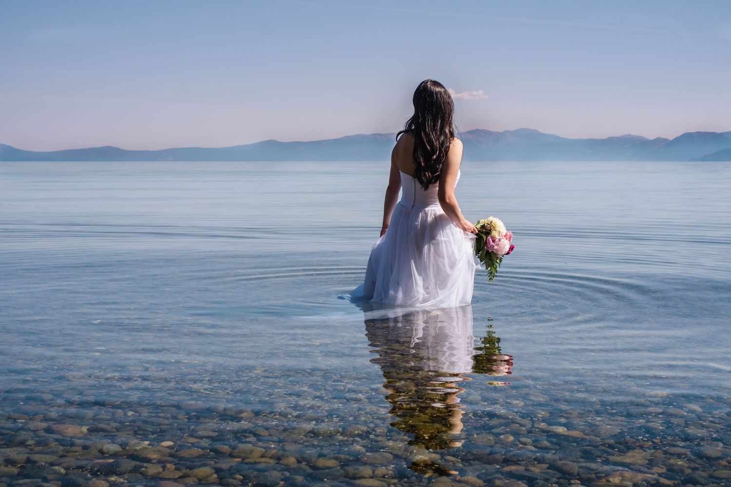 A bride in a white wedding dress stands in calm crystal clear waters of lake Tahoe California during her trash the dress bridal session