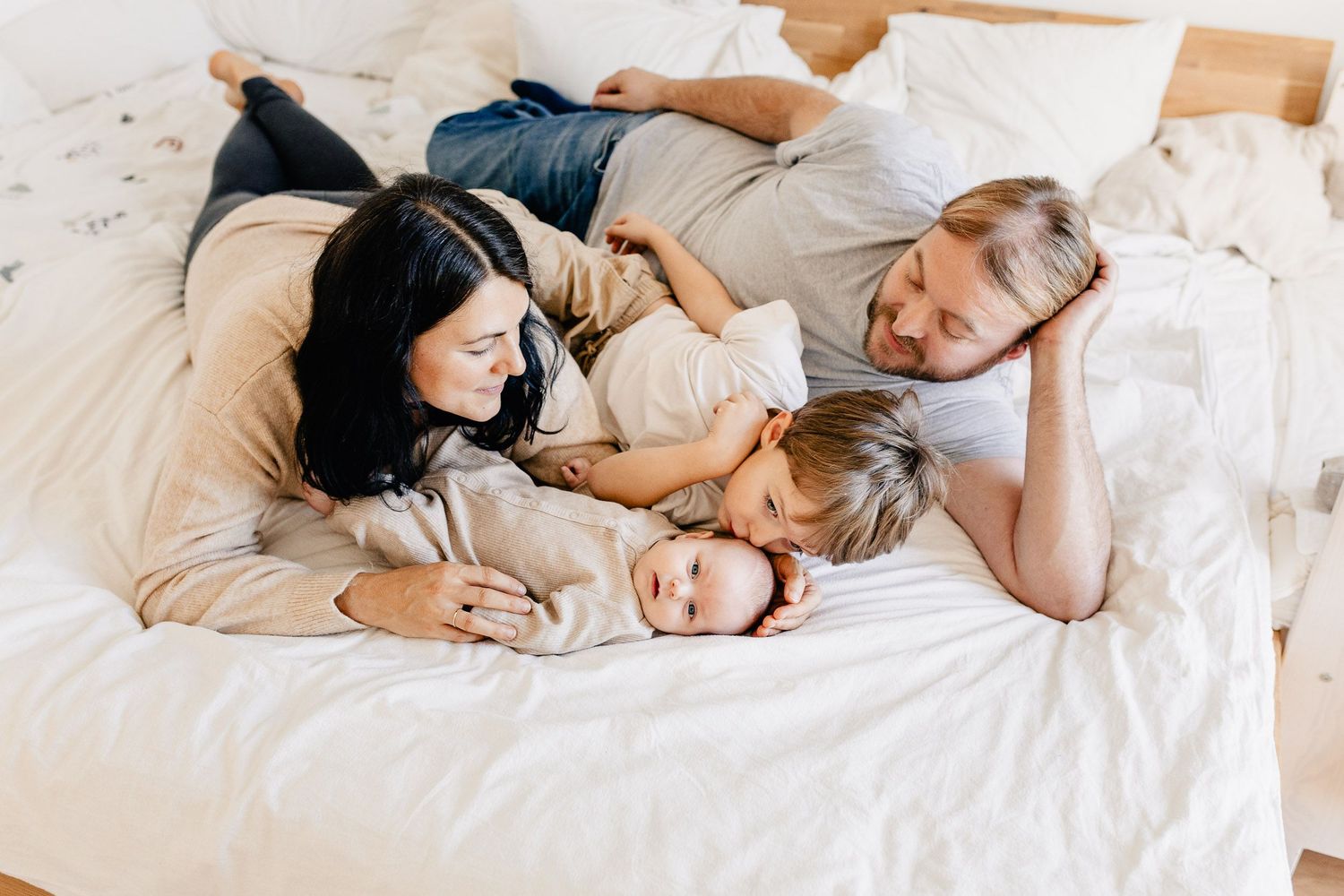 Familie kuschelt mit ihrem Baby zu Hause im Bett. Der Bruder gibt dem Baby einen Kuss.