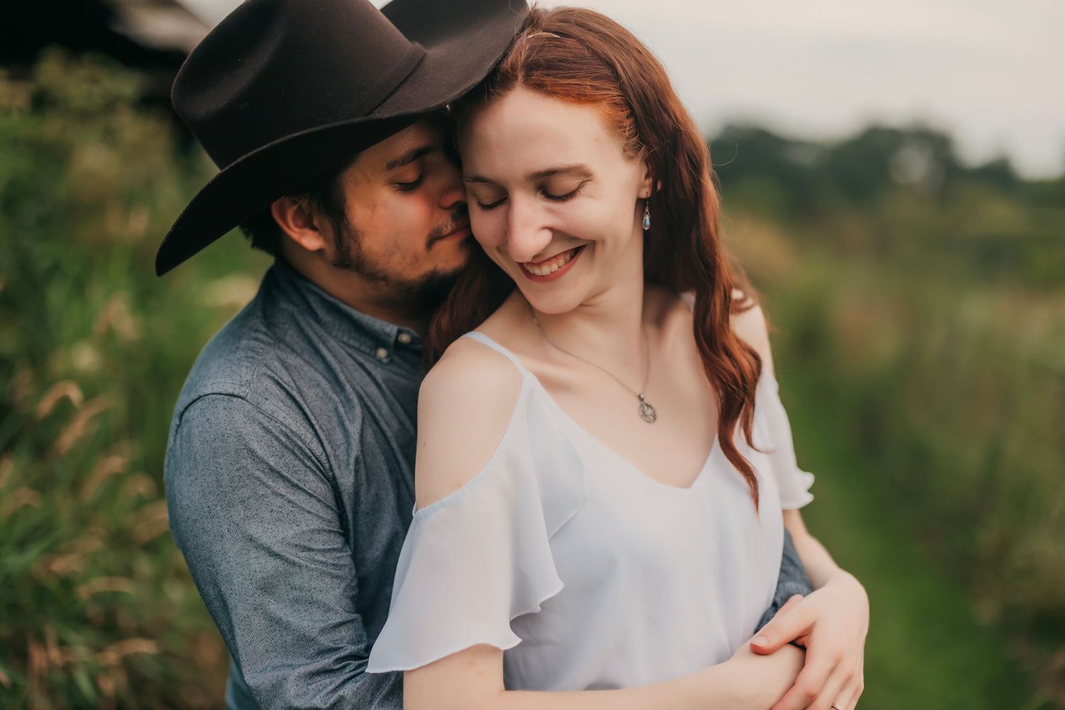 A tender romantic moment between a couple embracing in a sunlit meadow, the man wearing a dark hat and the woman in white.