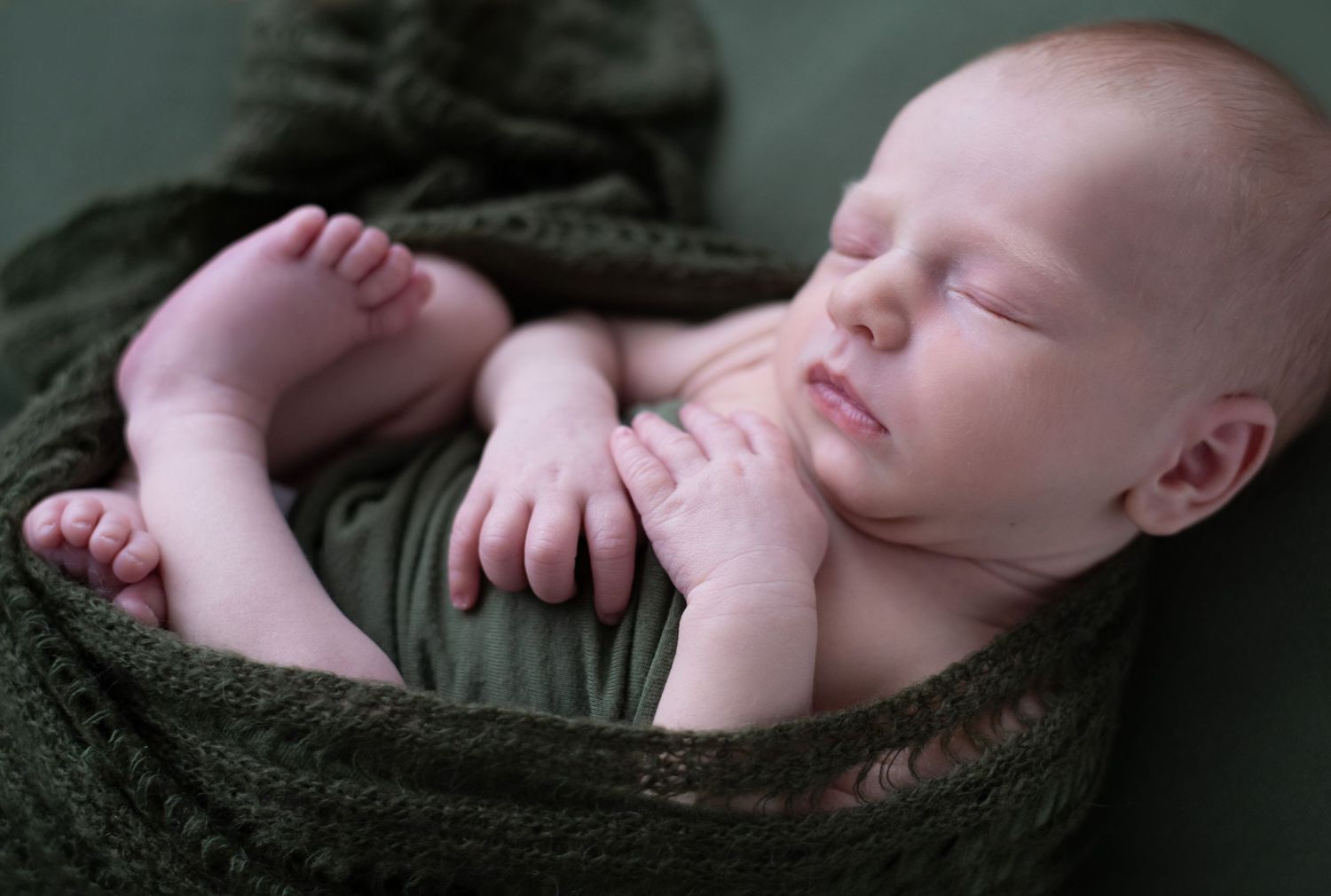 A peaceful sleeping newborn baby wrapped in a soft olive green blanket shows tiny pink toes and hands.
