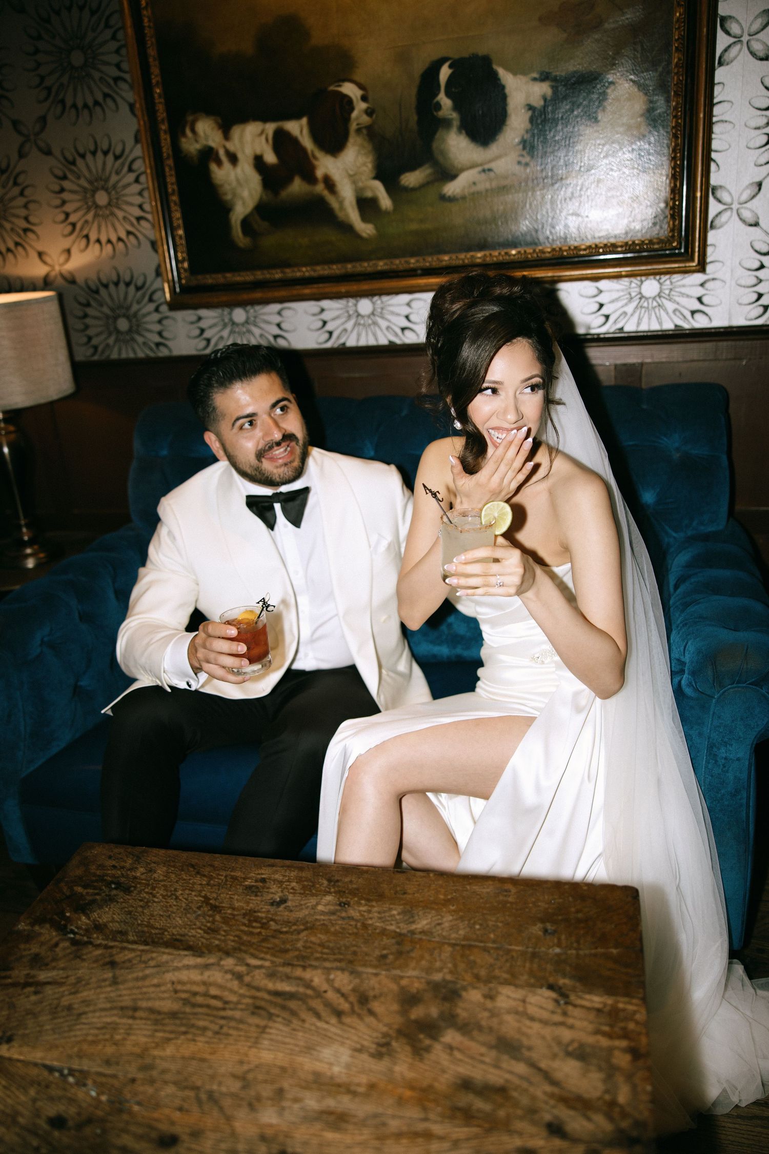 Bride and groom smiling with cocktails on a blue velvet sofa beneath a classic dog painting.