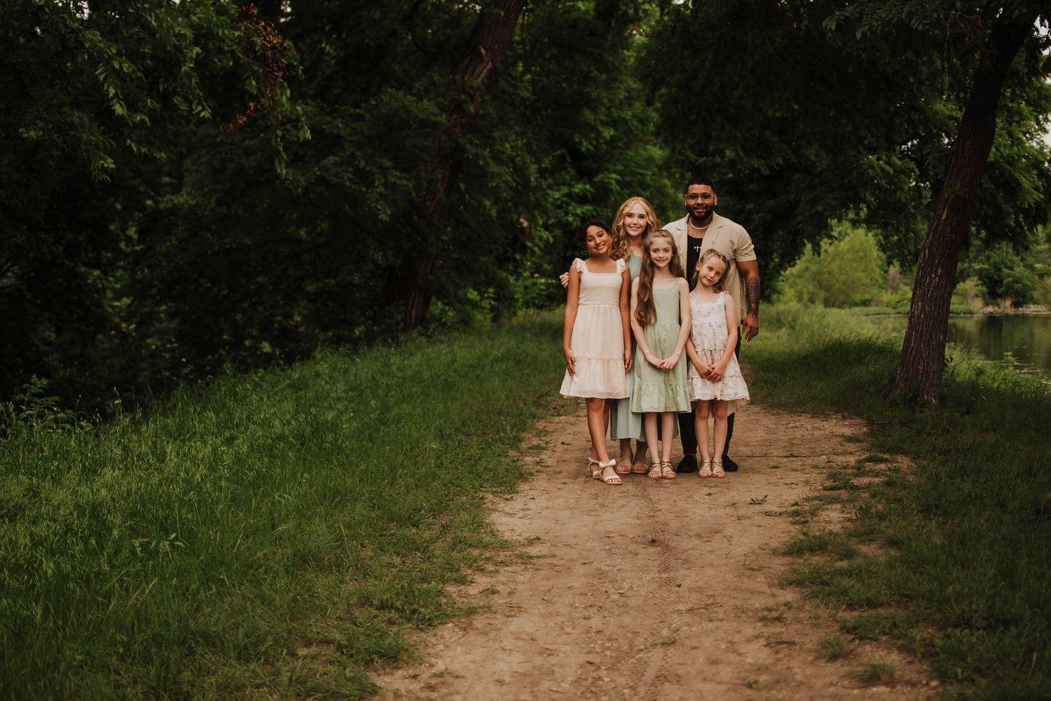 People in light colored dresses walking together on a dirt path through a green wooded area.