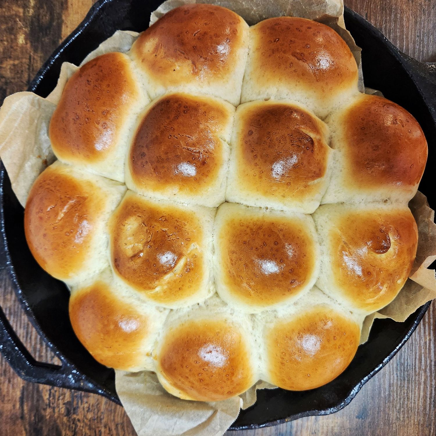Freshly baked golden brown dinner rolls arranged in a cast iron skillet with a glossy buttered top finish.