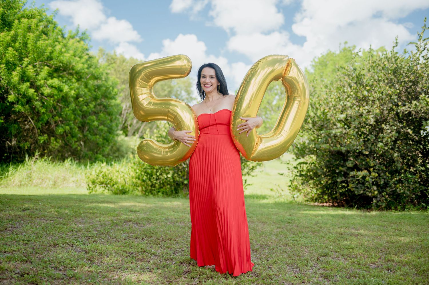 A person in a flowing red dress poses with golden number balloons 30 in a lush summer meadow.