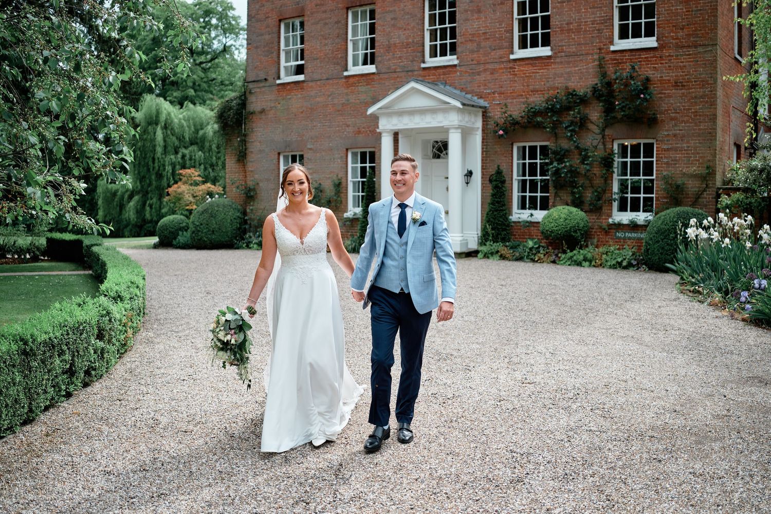 Wedding party walks down a gravel path beside a historic red brick mansion with classic Georgian windows.
