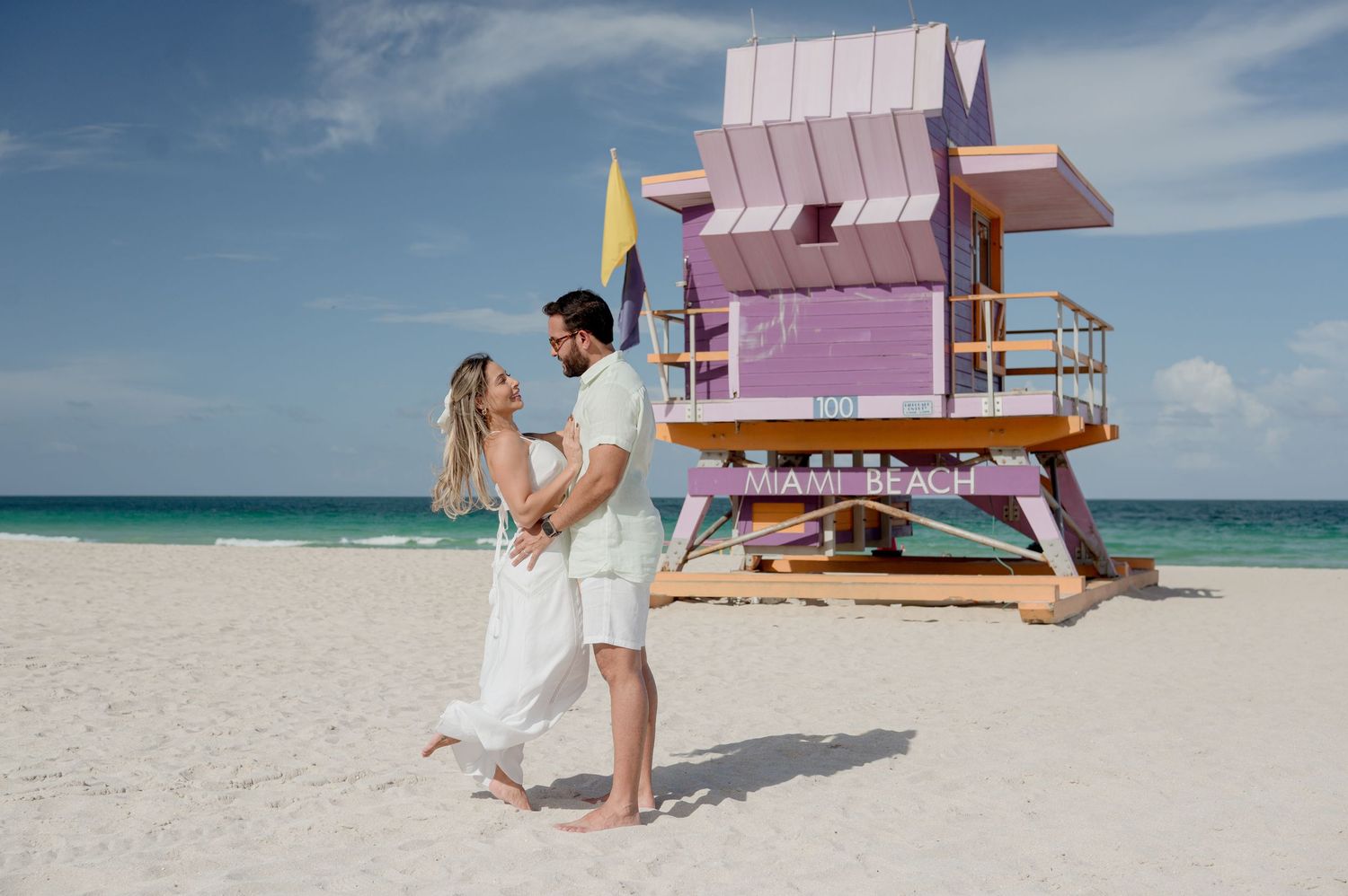 A couple stands near a pink lifeguard tower on a pristine Miami Beach with turquoise ocean waters and blue sky.
