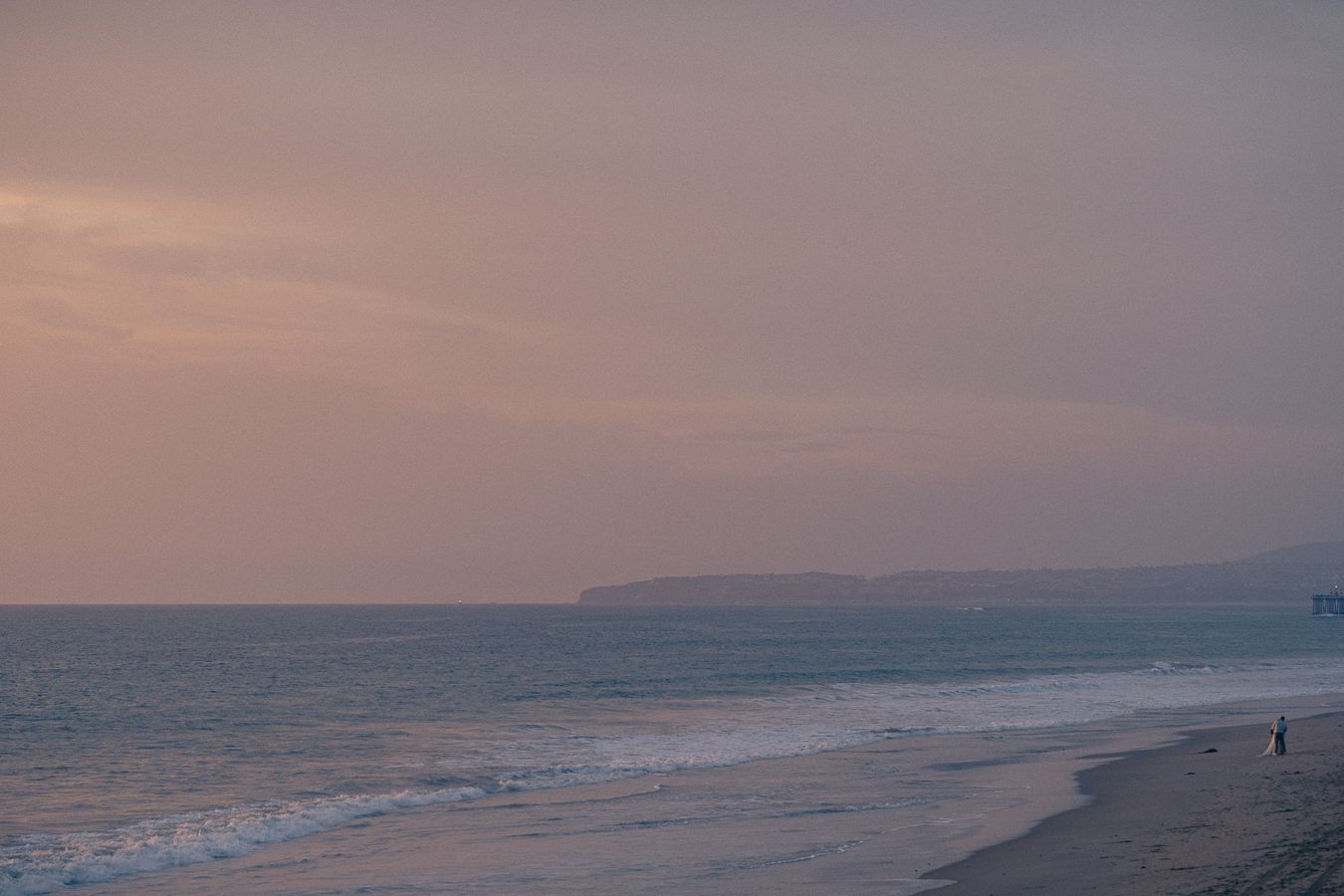 Scenic coastal landscape with waves crashing on beach during sunset with mountains in the distance.