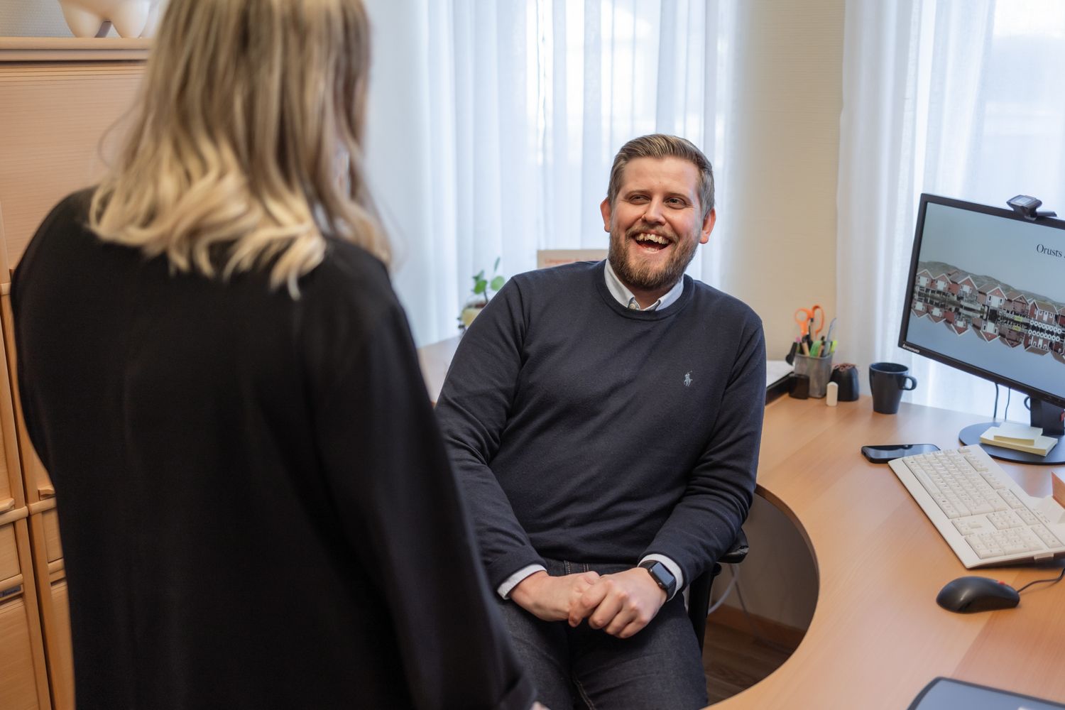 A person sits at a desk in a bright office environment, gesturing while engaged in conversation.