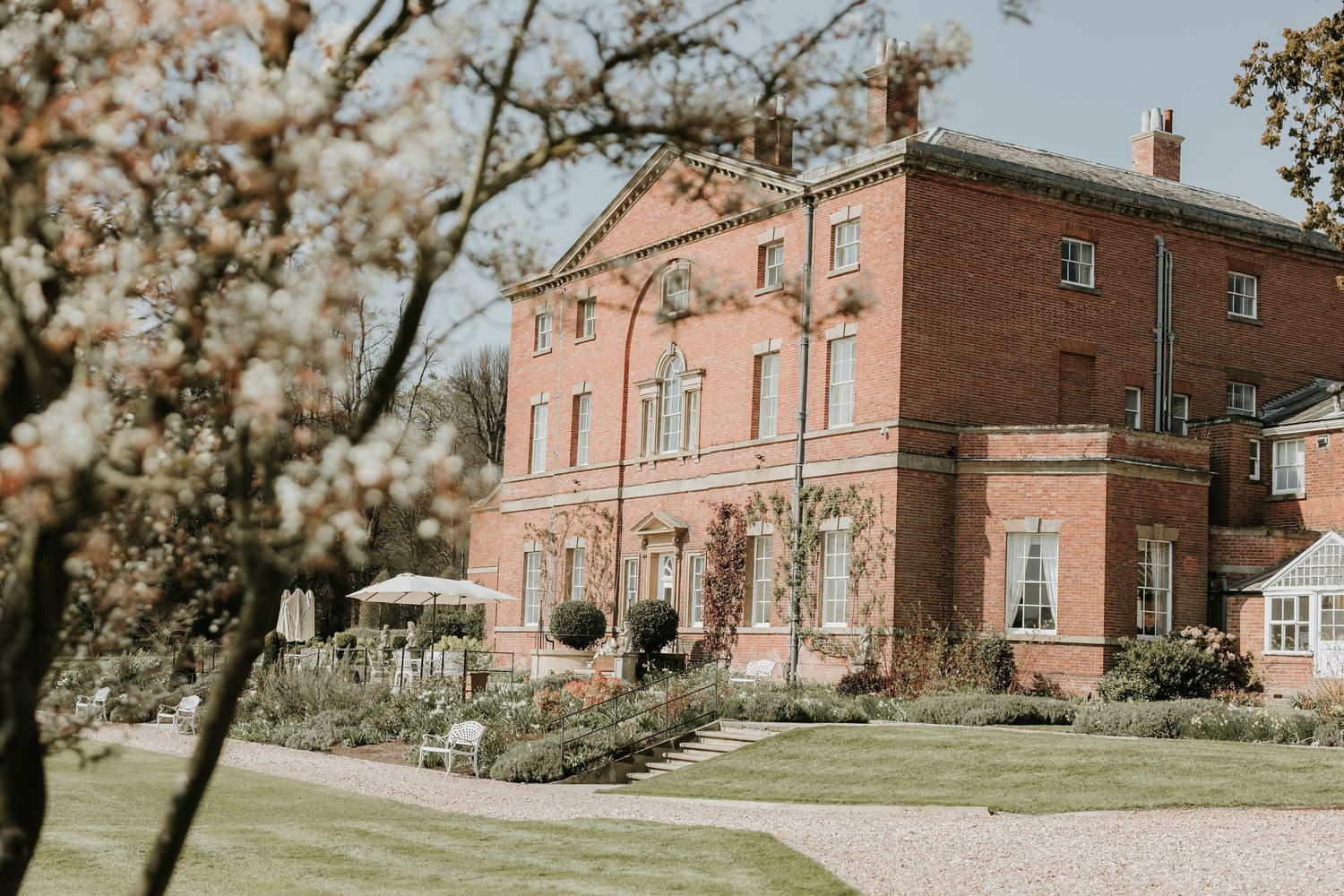Elegant brick mansion with white trim and columns set against blooming spring trees in soft natural lighting.